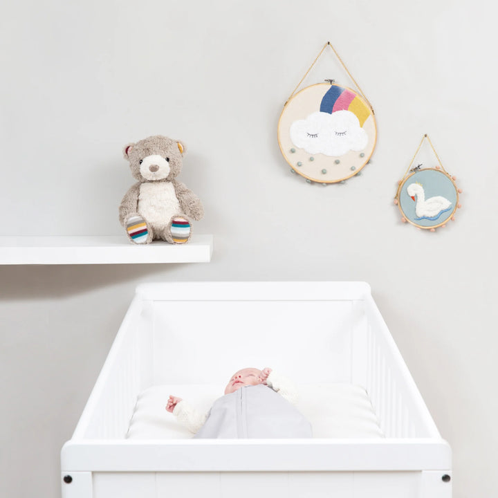 Zazu Bruno Baby Soother plush teddy bear displayed on nursery shelf beside baby sleeping peacefully in white cot, stylish and soothing sleep aid for newborns and nursery decor.