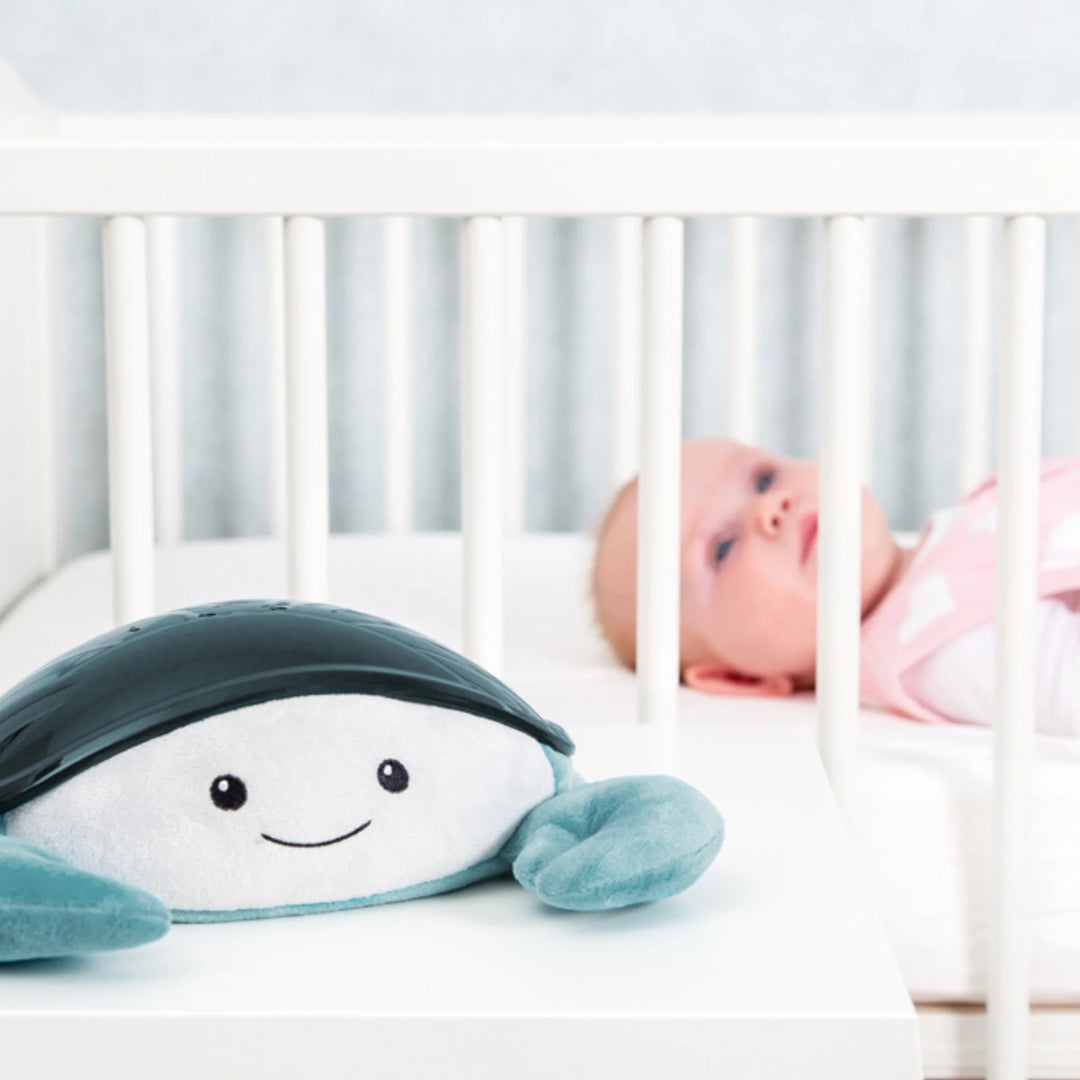 Zazu Cody Ocean Projector – plush blue crab nightlight placed beside a baby in a cot, creating a soothing and comforting sleep environment with calming ocean lights and melodies.