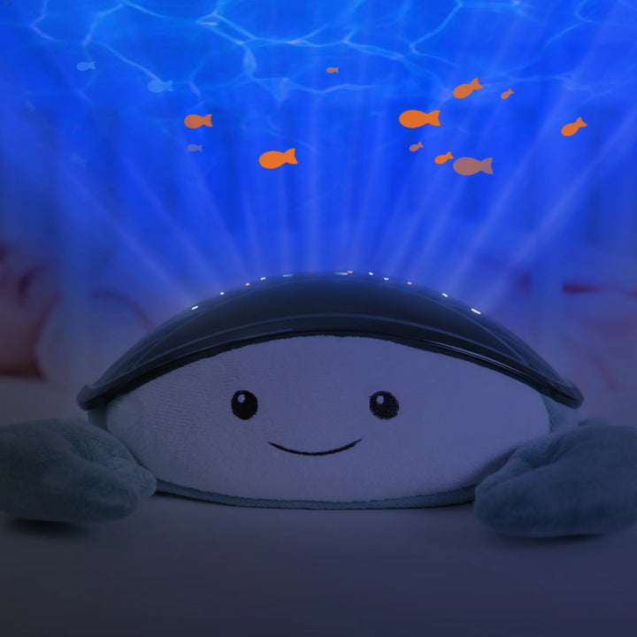 Zazu Cody Ocean Projector creating soothing underwater light effects beside a baby in a crib, projecting moving fish and calming blue waves to help babies relax and fall asleep peacefully.