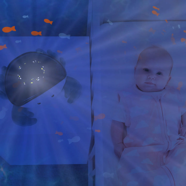 Zazu Cody Ocean Projector creating soothing underwater light effects beside a baby in a crib, projecting moving fish and calming blue waves to help babies relax and fall asleep peacefully.