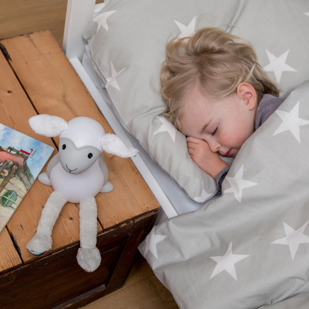 Toddler sleeping peacefully beside ZAZU Fin the Sheep Soft Toy Nightlight on bedside table — gentle glowing nightlight for babies and toddlers providing comfort and security during sleep. Ideal bedtime companion and sleep training aid for children’s nighttime routines.