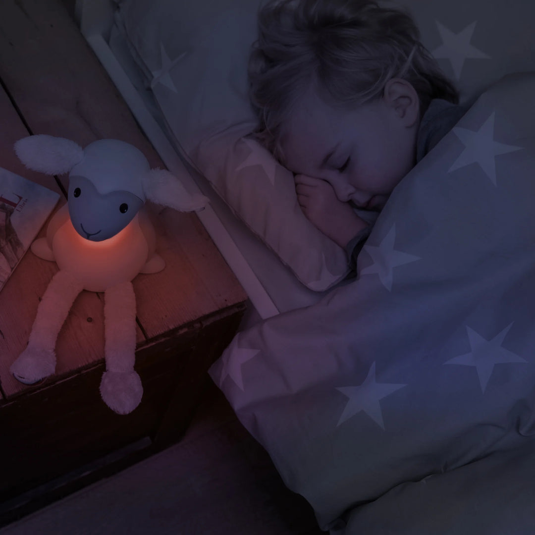 oddler sleeping peacefully beside ZAZU Fin the Sheep Nightlight glowing with soft red light — soothing bedtime lamp for children promoting restful sleep and comfort in nursery or kids’ bedroom.
