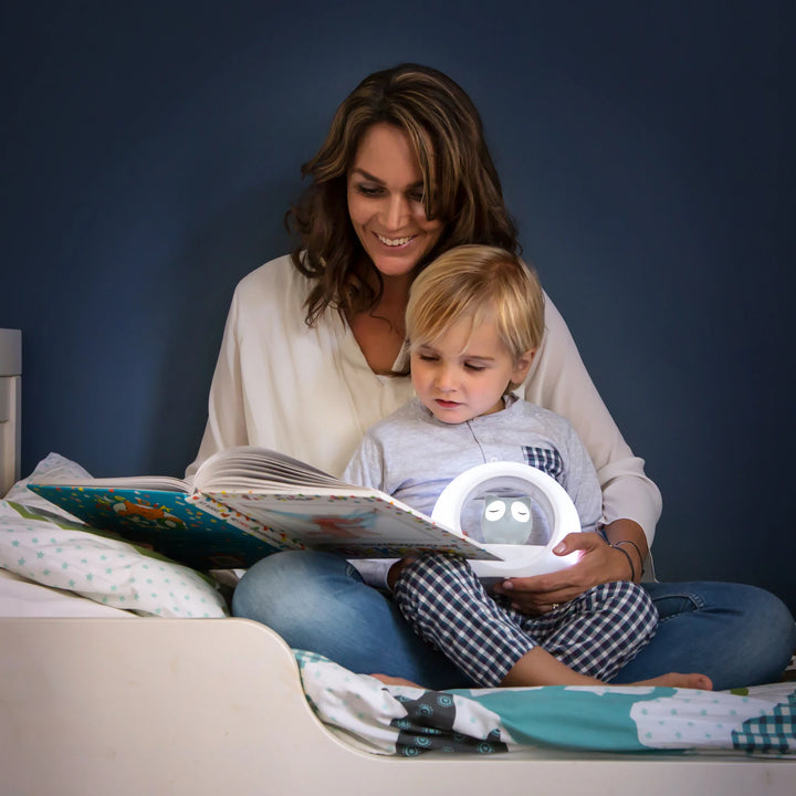 Zazu Lou Nightlight – glowing owl nightlight on bedside table beside an awake child, providing soft adjustable light and cry sensor comfort to help children feel safe at bedtime.