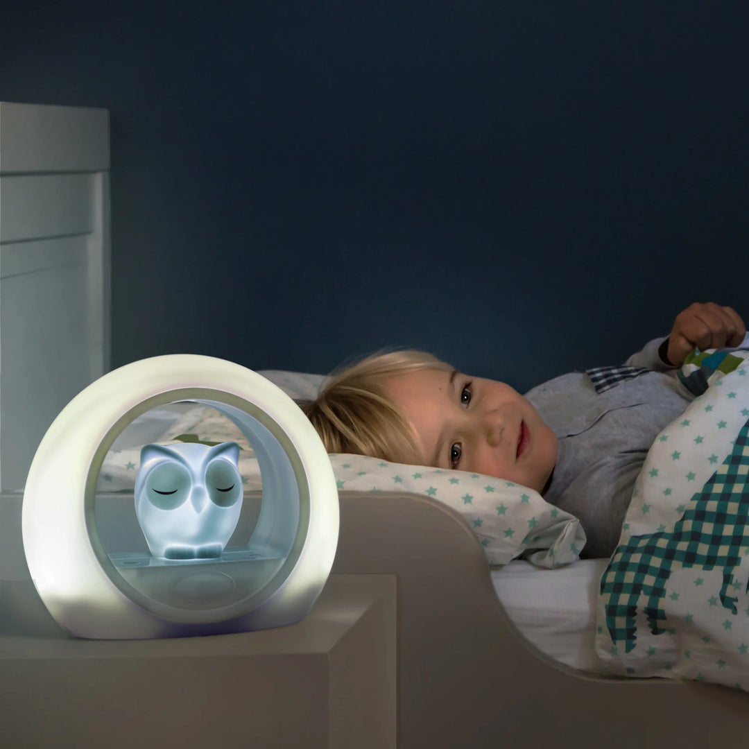 Zazu Lou Nightlight – glowing owl nightlight on bedside table beside an awake child, providing soft adjustable light and cry sensor comfort to help children feel safe at bedtime.