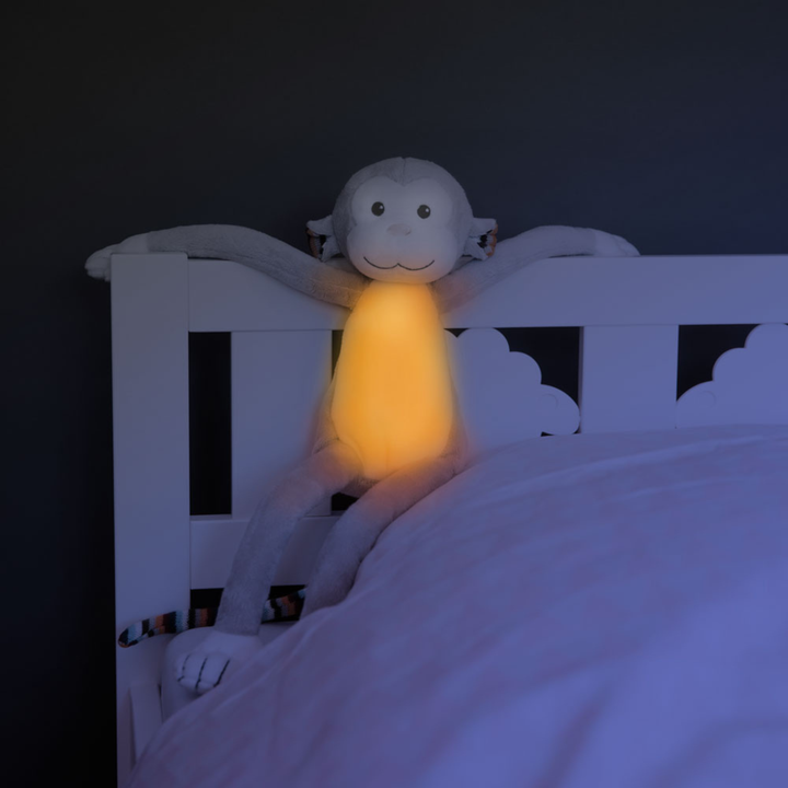 ZAZU Max the Monkey Nightlight glowing softly on a child’s bed frame at night, showing its warm amber belly light. Baby sleep aid plush toy with soothing illumination designed to comfort children and promote peaceful sleep routines.