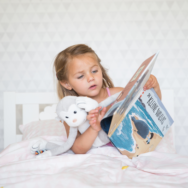 Little girl reading a bedtime story in bed while cuddling the ZAZU Max the Monkey Nightlight plush toy. Soft toy nightlight for children providing warm light and comfort during bedtime routines — perfect sleep aid for toddlers.