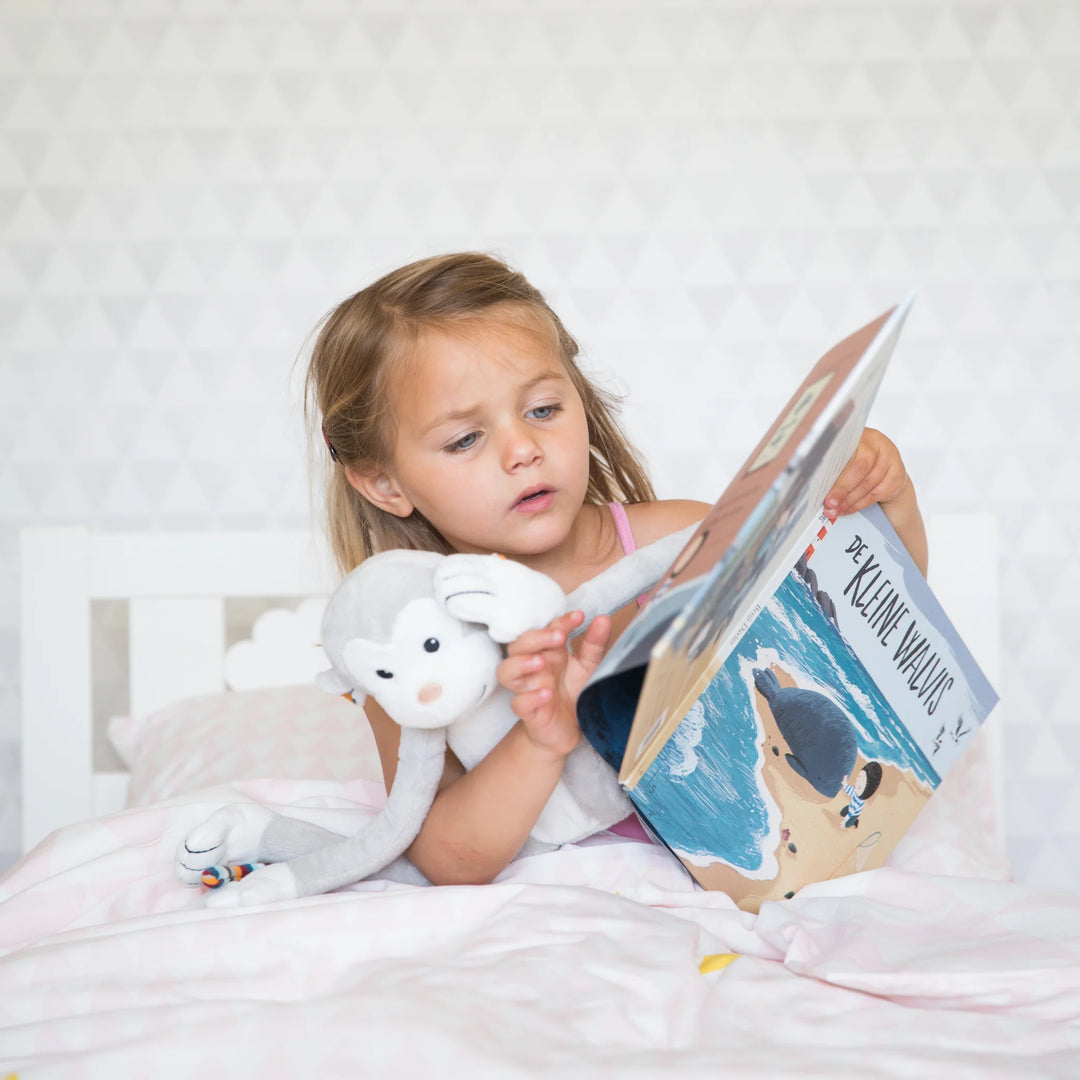 Child reading a bedtime story in bed while cuddling the Zazu Max soft toy nightlight plush monkey, showing comforting bedtime use of the glowing nightlight for toddlers.