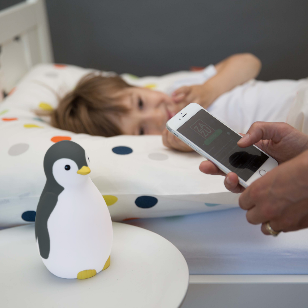 Parent adjusting Zazu Pam the Penguin Sleep Trainer settings on smartphone beside a child in bed – smart children’s sleep training clock and night light with app control, color-changing lights, and soothing sounds. Helps toddlers learn healthy sleep and wake routines.
