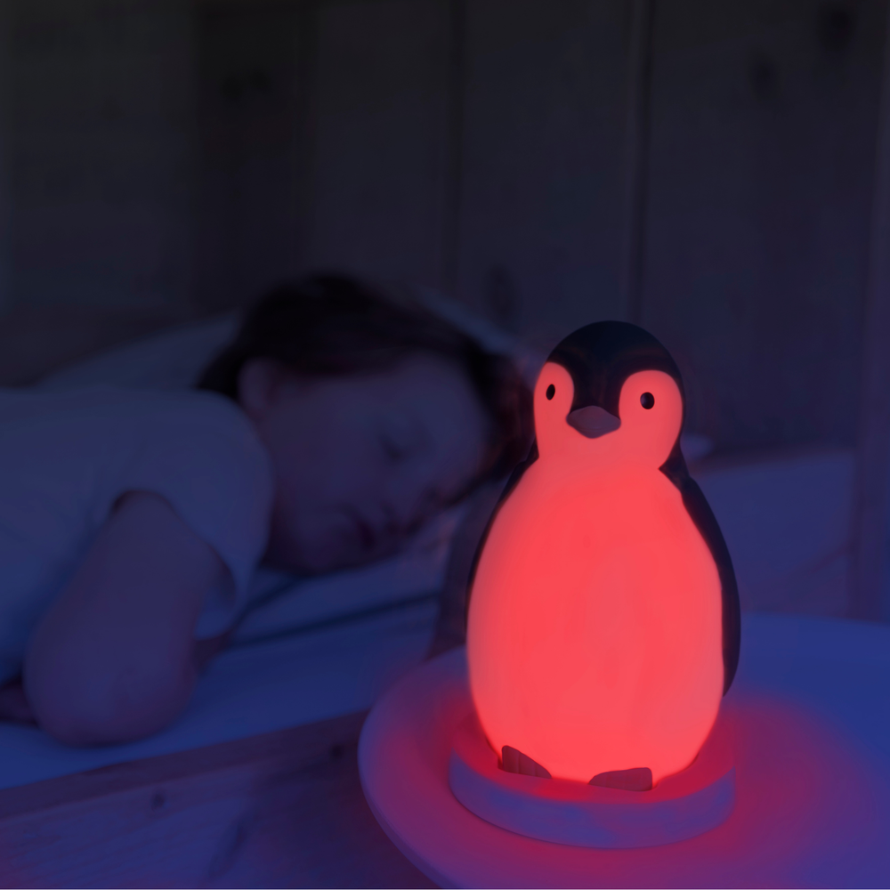 “Zazu Pam the Penguin Sleep Trainer glowing red night light beside a sleeping child – bedtime sleep aid that teaches children when to sleep and wake with color-changing lights and soothing sounds. Ideal night light and sleep training clock for toddlers and preschoolers.