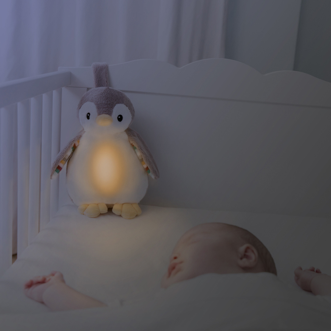 ZAZU Phoebe penguin baby soother glowing softly in crib beside sleeping baby. Plush penguin nightlight and sound machine emits warm light and soothing sounds to comfort infants and promote restful sleep at bedtime