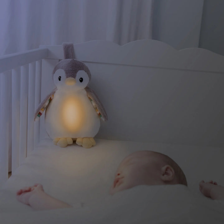 ZAZU Phoebe penguin baby soother glowing with warm nightlight inside a crib beside a sleeping baby.