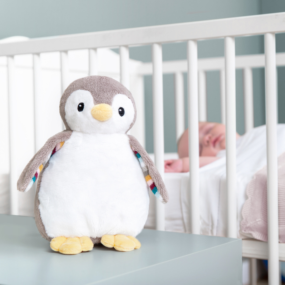 ZAZU Phoebe Baby Soother penguin plush toy beside baby crib – adorable grey and white penguin nightlight and sound machine for newborns, helping soothe and calm babies for better sleep.
