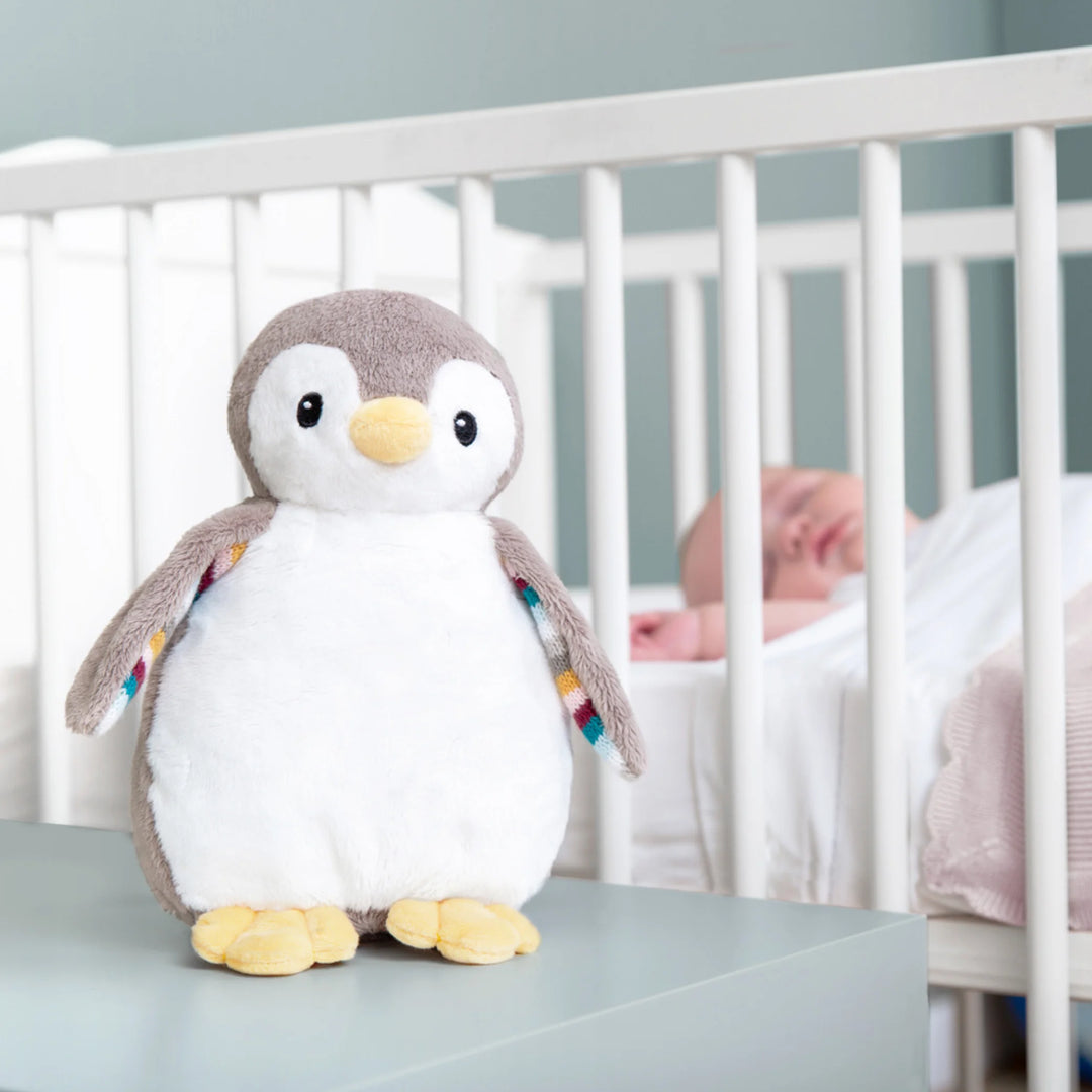 ZAZU Phoebe penguin baby soother plush displayed on a nursery table with a sleeping baby in a crib in the background.