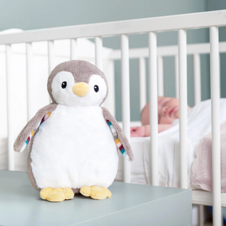 ZAZU Phoebe penguin baby soother plush displayed on a nursery table with a sleeping baby in a crib in the background.