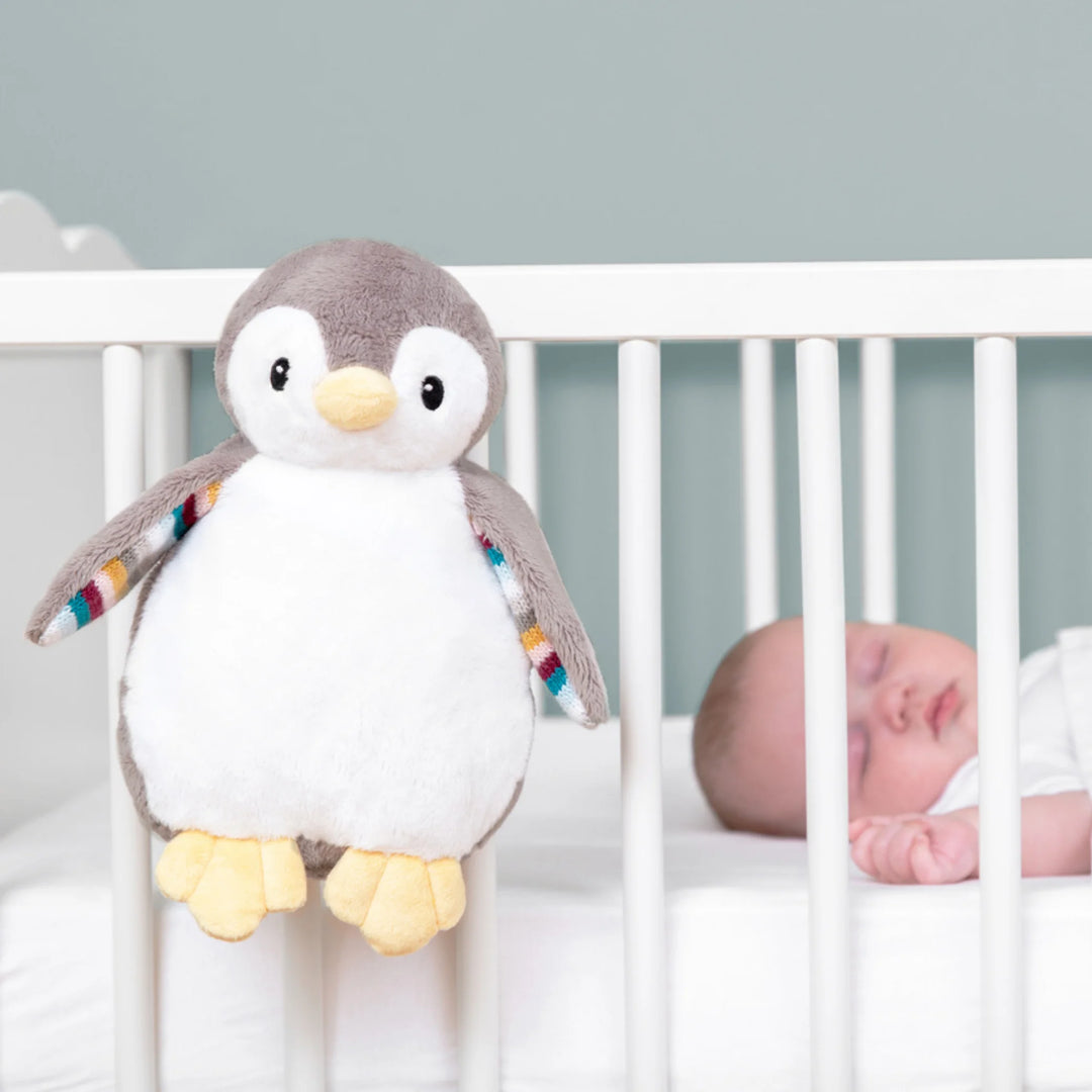 ZAZU Phoebe penguin baby soother attached to a crib rail beside a sleeping baby in a nursery.