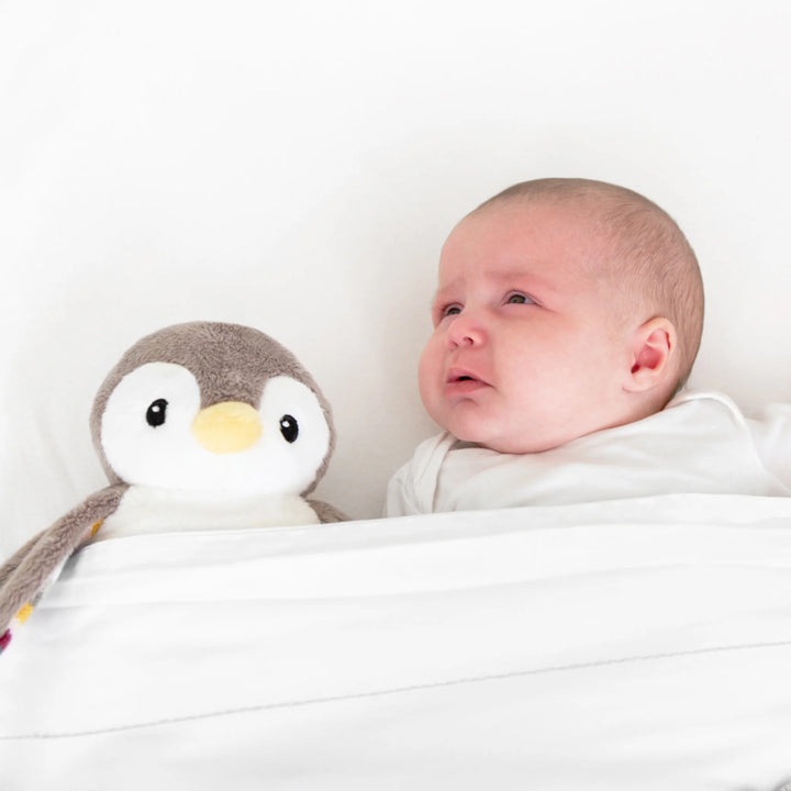Crying baby lying in bed beside the ZAZU Phoebe penguin plush baby soother designed to comfort infants.