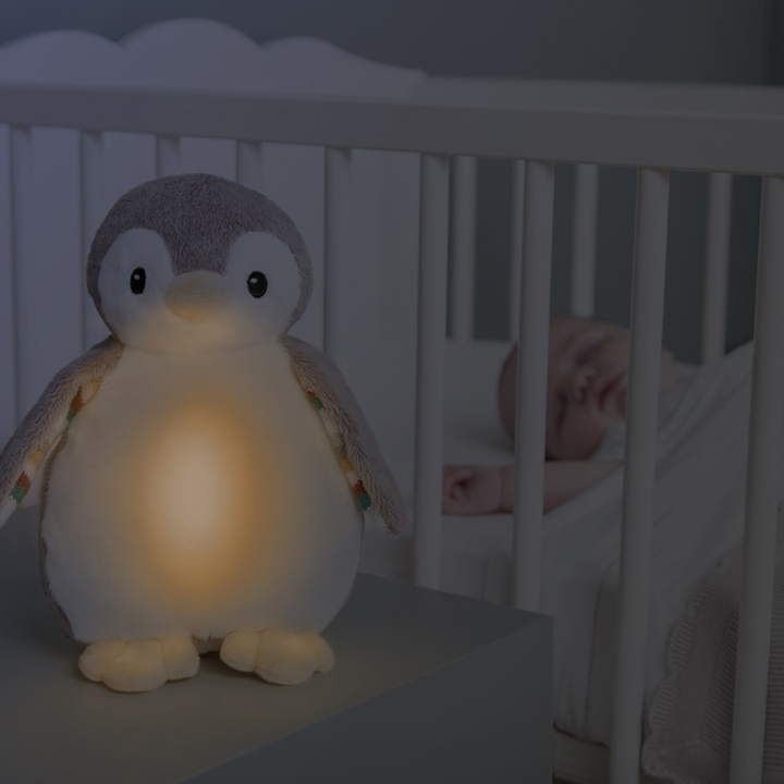 ZAZU Phoebe Baby Soother penguin nightlight glowing softly beside a sleeping baby in crib – soothing bedtime toy with heartbeat, white noise, and lullaby sounds for newborn sleep comfort.