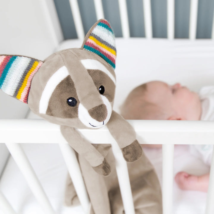 ZAZU Robin raccoon baby comforter hanging on a white crib beside a sleeping newborn, soft plush security blanket used as a soothing sleep aid.