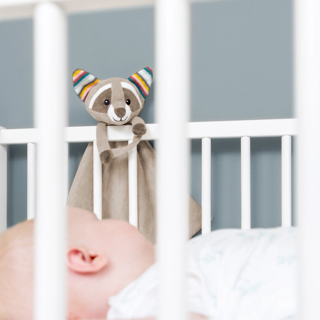 ZAZU Robin raccoon baby comforter hanging on a white crib while a newborn sleeps, soft plush security blanket used as a soothing sleep aid in the nursery.