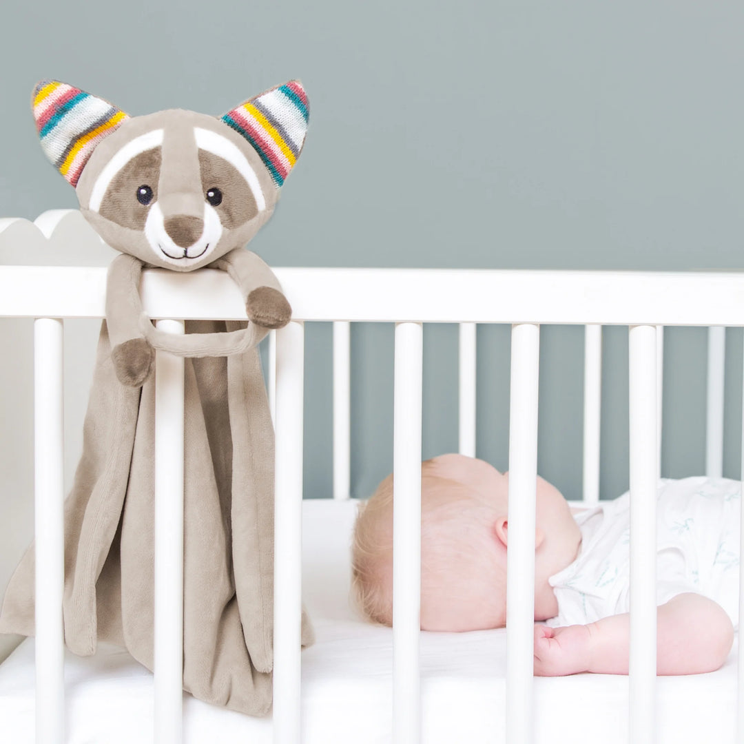ZAZU Robin raccoon baby comforter hanging on a white crib beside a sleeping newborn, soft plush security blanket providing soothing comfort in the nursery
