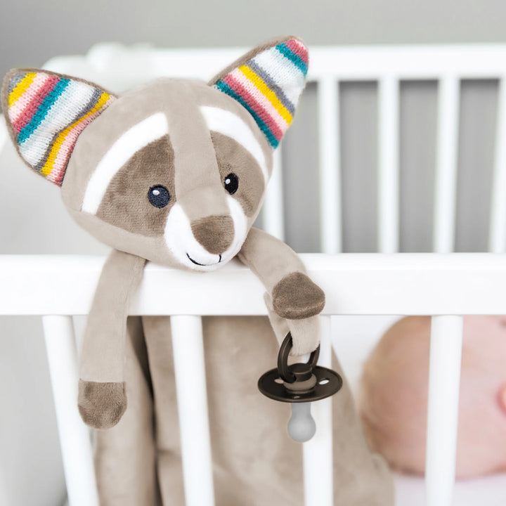 ZAZU Robin raccoon baby comforter hanging on a white crib, holding a pacifier beside a sleeping newborn - plush security blanket and soothing sleep aid for babies.