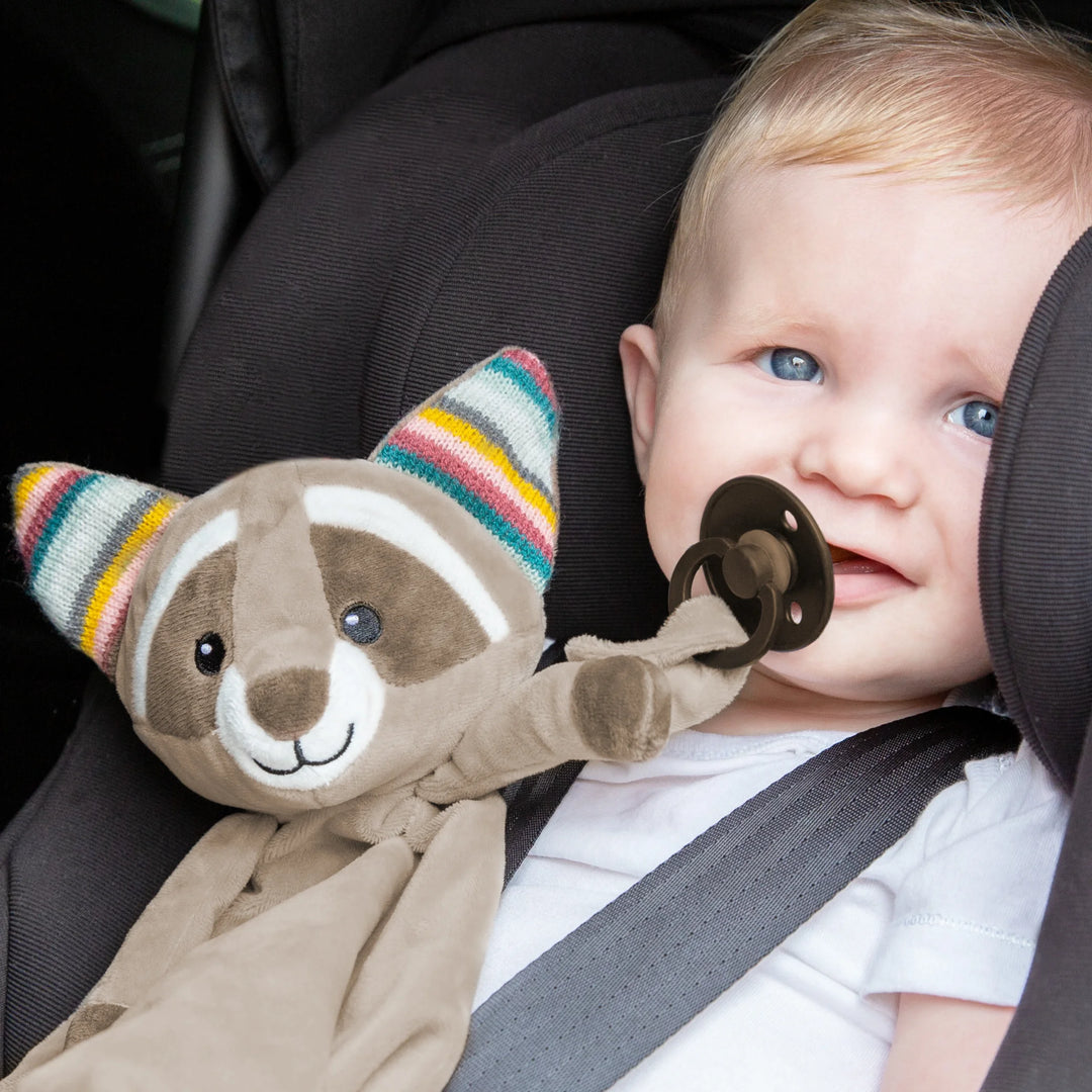 Baby sitting in a car seat holding the ZAZU Robin raccoon baby comforter with pacifier attachment — soft plush security blanket designed to soothe and comfort infants on the go