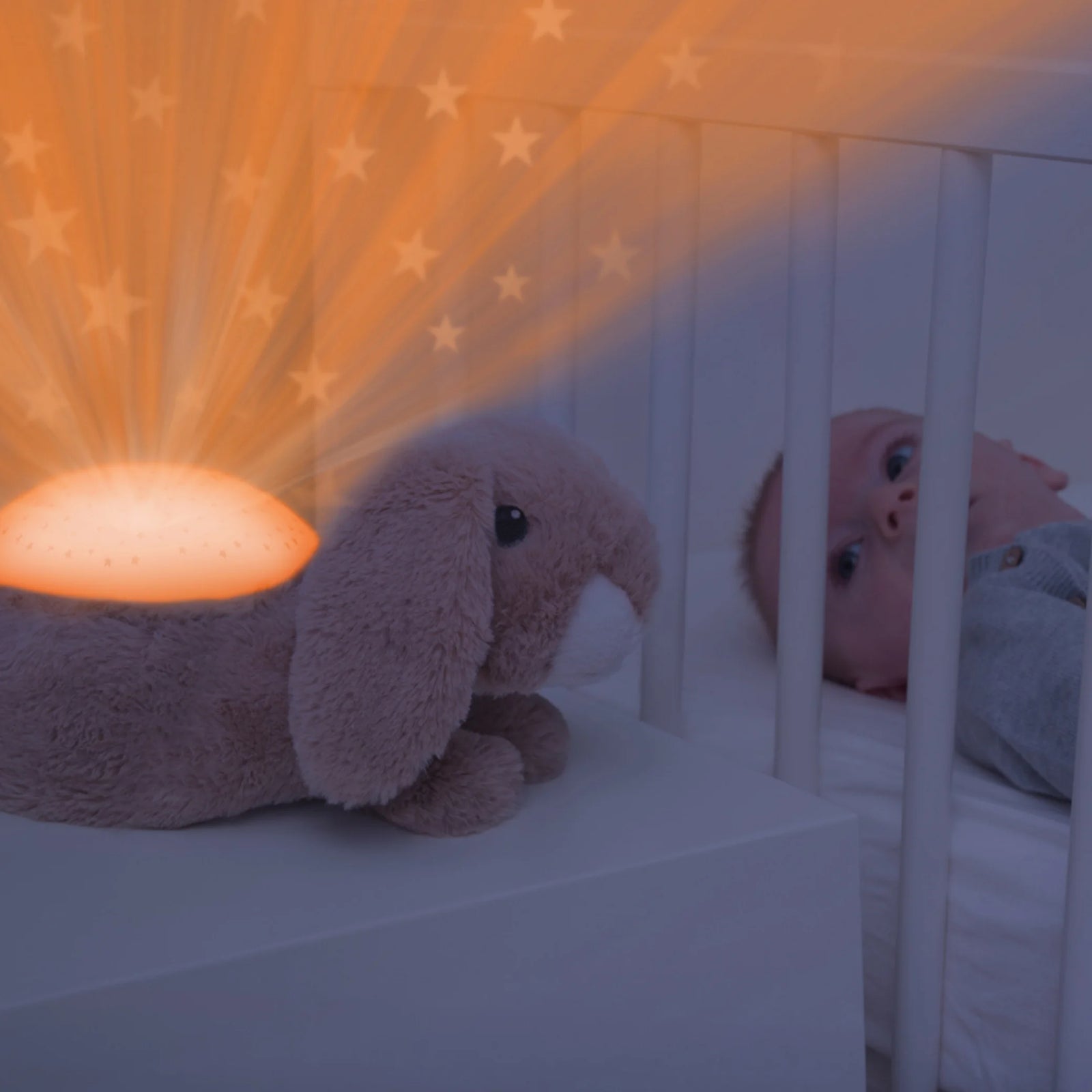 “Baby lying in crib watching the Zazu Ruby Star Projector bunny nightlight projecting warm orange stars onto the nursery wall, soothing sleep aid with gentle light and melodies