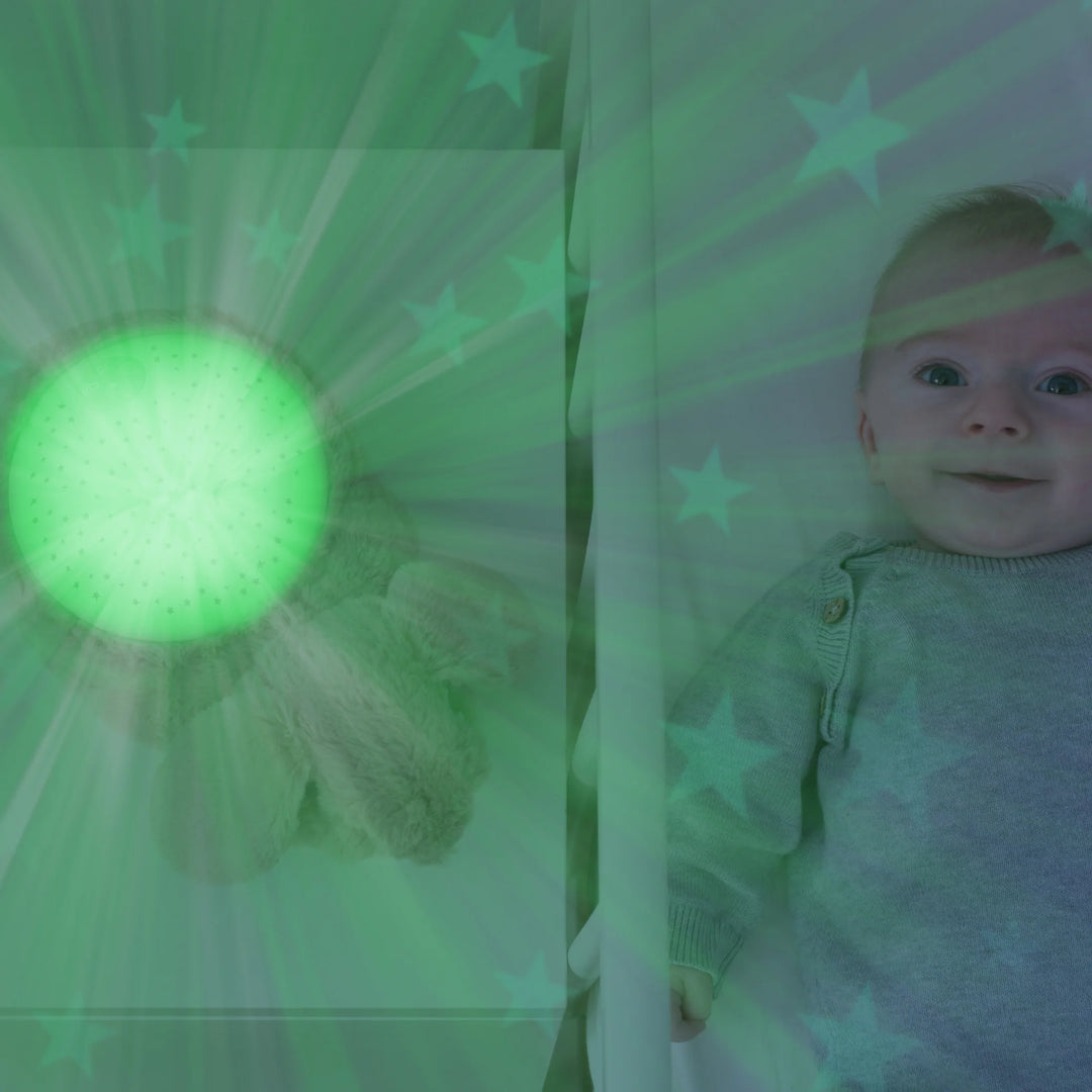 Baby smiling in crib beside ZAZU Ruby the Rabbit Star Projector emitting soft green light and star pattern on walls, soothing nursery night light with music and cry sensor for better baby sleep routines.