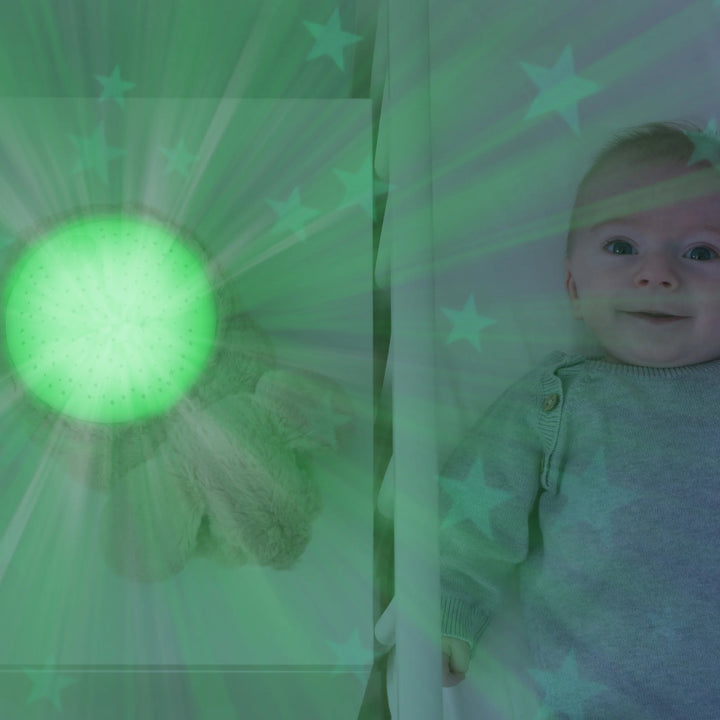 Baby smiling in crib beside ZAZU Ruby the Rabbit Star Projector emitting soft green light and star pattern on walls, soothing nursery night light with music and cry sensor for better baby sleep routines.