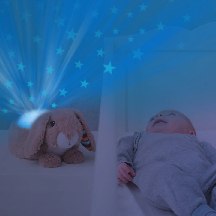 Baby lying in crib smiling while ZAZU Ruby the Rabbit Star Projector emits soothing blue starry light patterns on the wall and ceiling, creating a calming nursery atmosphere with gentle night light projection for better baby sleep.