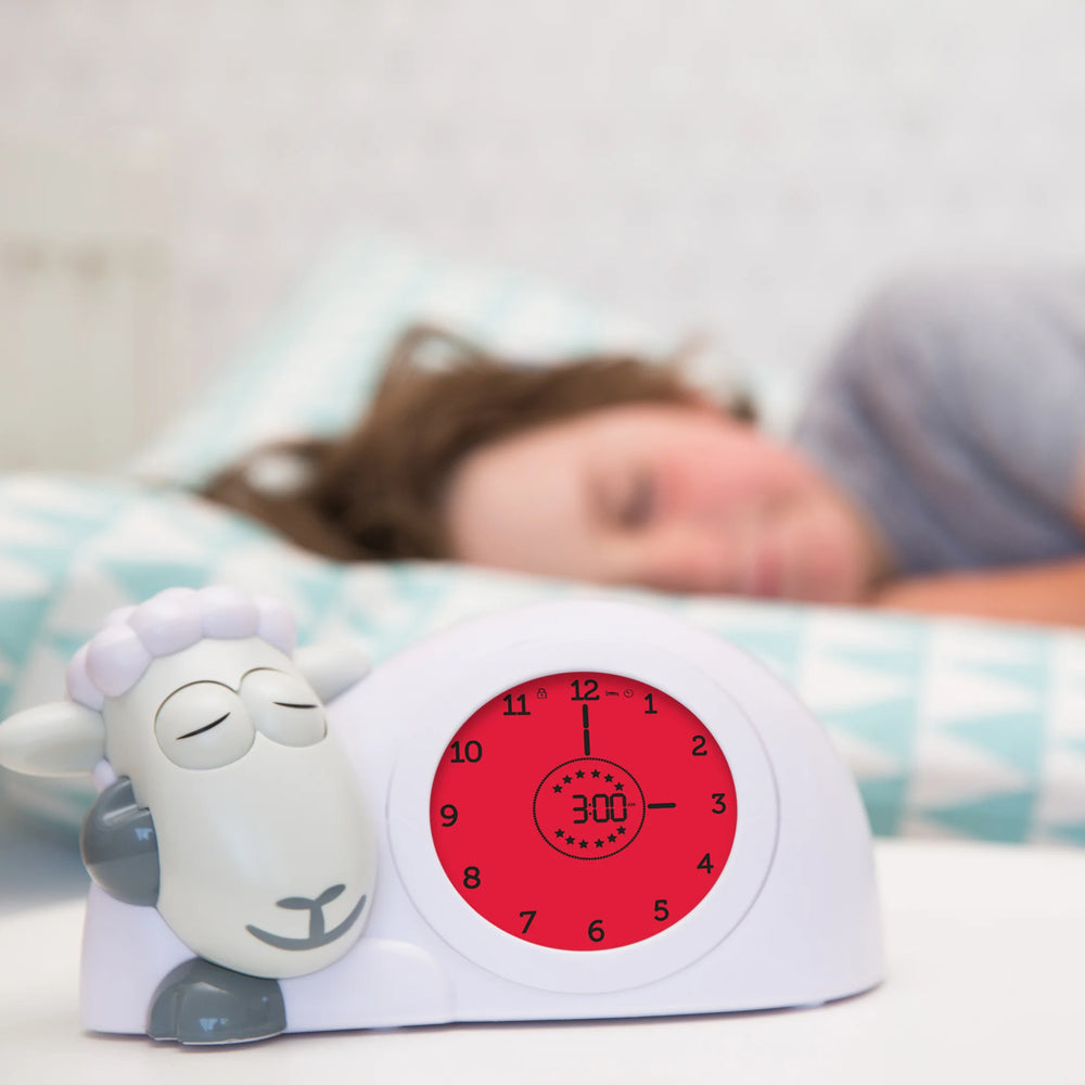 Zazu Sam the Sheep Sleep Trainer with red night mode light beside a sleeping child – children’s sleep training clock that teaches bedtime and wake-up routines using color-changing screens and visual cues. Perfect toddler sleep aid and kids’ alarm clock.