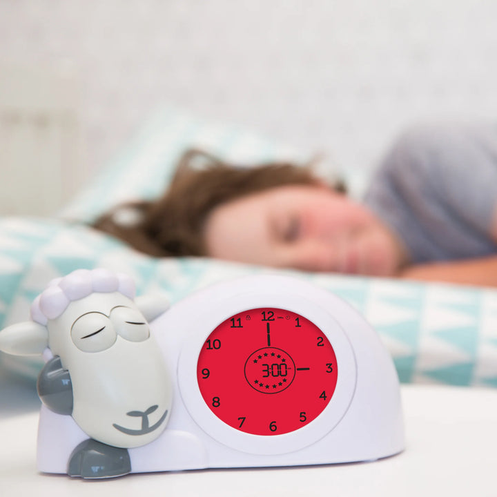 Zazu Sam the Sheep Sleep Trainer with red night mode light beside a sleeping child – children’s sleep training clock that teaches bedtime and wake-up routines using color-changing screens and visual cues. Perfect toddler sleep aid and kids’ alarm clock.