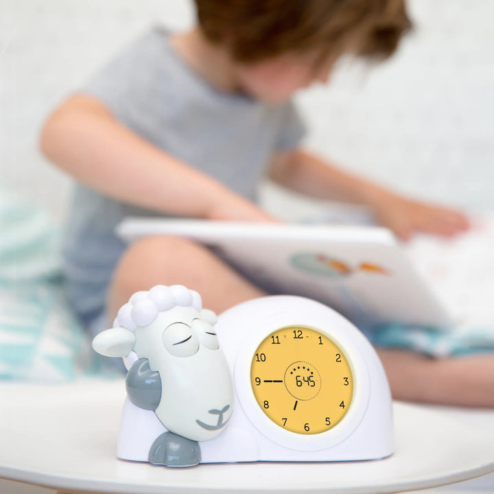 “Zazu Sam the Sheep Sleep Trainer showing yellow ‘almost wake-up’ light beside a child reading in bed – children’s sleep training clock that teaches kids when to stay in bed or wake up using color-changing lights. Ideal bedtime routine clock and toddler sleep aid.