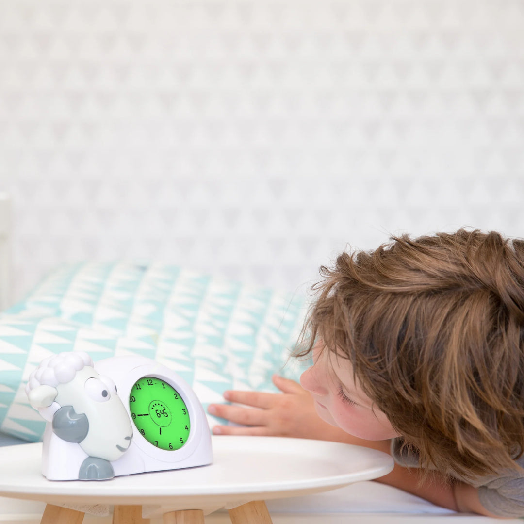 Child waking up and looking at the ZAZU Sam the Sheep sleep trainer clock in green wake-up mode, showing the ‘OK to get up’ display on a bedside table in a bright bedroom.