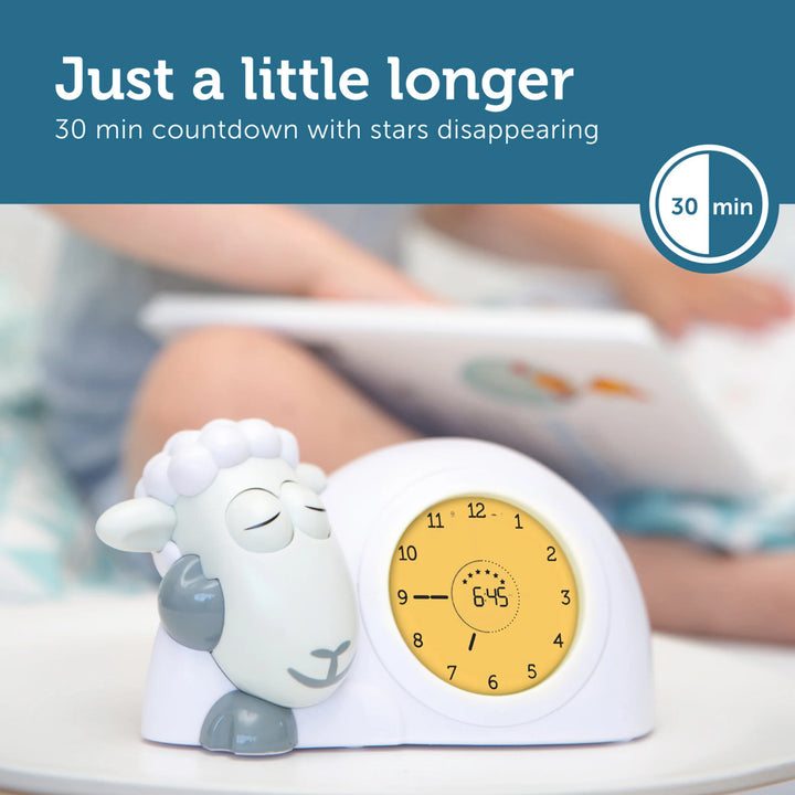 ZAZU Sam the Sheep sleep trainer clock in yellow countdown mode with sleepy sheep character, shown beside a child reading, demonstrating the 30-minute star countdown feature.