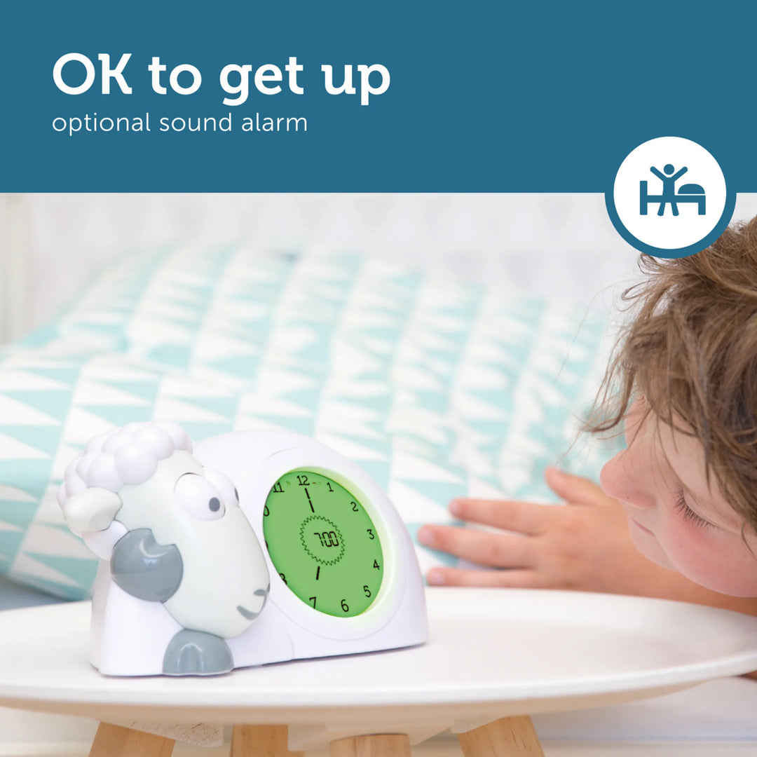 ZAZU Sam the Sheep sleep trainer clock in green wake-up mode, shown beside a child in bed, highlighting the ‘OK to get up’ feature with optional sound alarm.