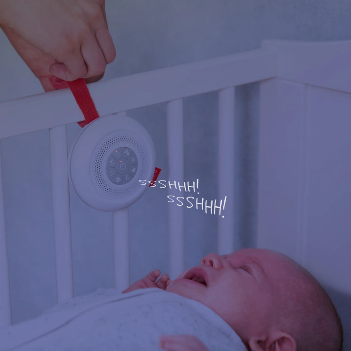 Crying baby in crib soothed by the ZAZU Suzy Portable Baby Soother emitting “shhh” white noise sounds. The sound machine is attached to the cot with a red strap, helping calm and comfort newborns to sleep. Ideal portable baby sleep aid with shushing sound feature for peaceful naps and bedtime routines.