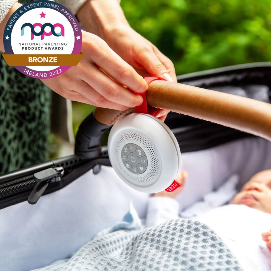 Close-up of a parent attaching the award-winning ZAZU Suzy Portable Baby Soother to a stroller handle beside a sleeping baby. The white noise machine features multiple soothing sounds and a secure red strap. Awarded Bronze in the National Parenting Product Awards Ireland 2022. Perfect for calming newborns on the go.