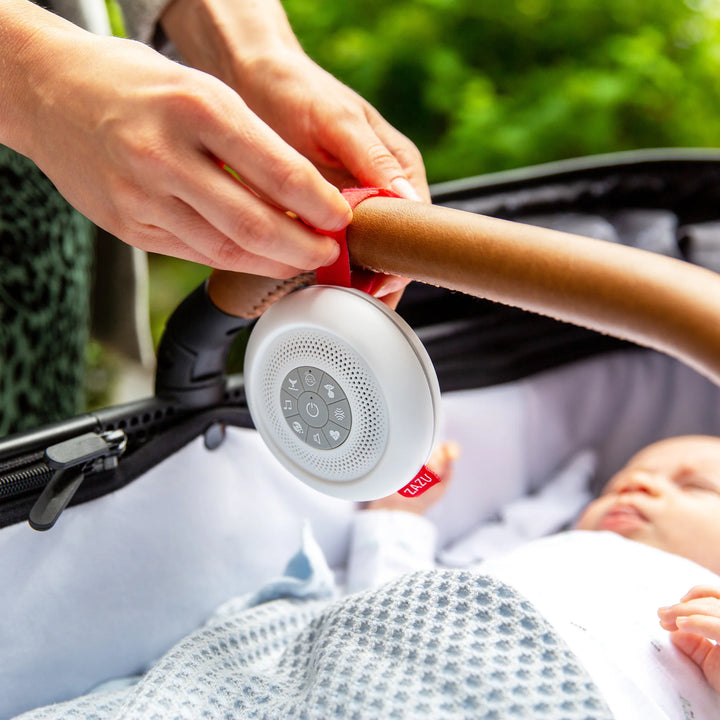 Parent attaching the ZAZU Suzy Portable Baby Soother with a red strap to a stroller handle while a baby rests peacefully inside. The white noise machine plays soothing sounds to calm babies on the go, making it the perfect travel sleep aid for prams, car seats, and cribs.