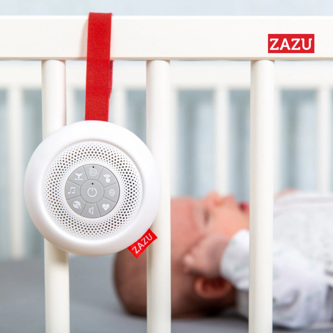 ZAZU Suzy portable baby soother attached to a white crib with a newborn lying inside, featuring soothing sound buttons and a red hanging strap