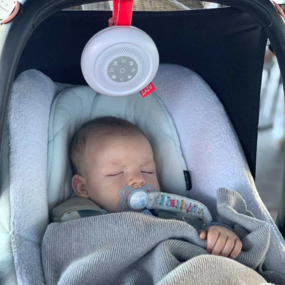 Sleeping baby in car seat soothed by the ZAZU Suzy Portable Baby Soother attached above with a red strap. The white noise machine plays calming sounds to help infants rest peacefully at home or on the go. Perfect travel sleep aid for newborns and parents seeking portable sleep solutions