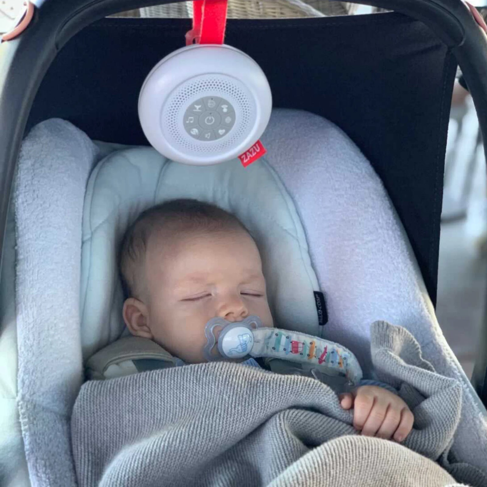 ZAZU Suzy portable baby sleep soother hanging from a car seat handle, playing calming sounds for a sleeping baby wrapped in a blanket.