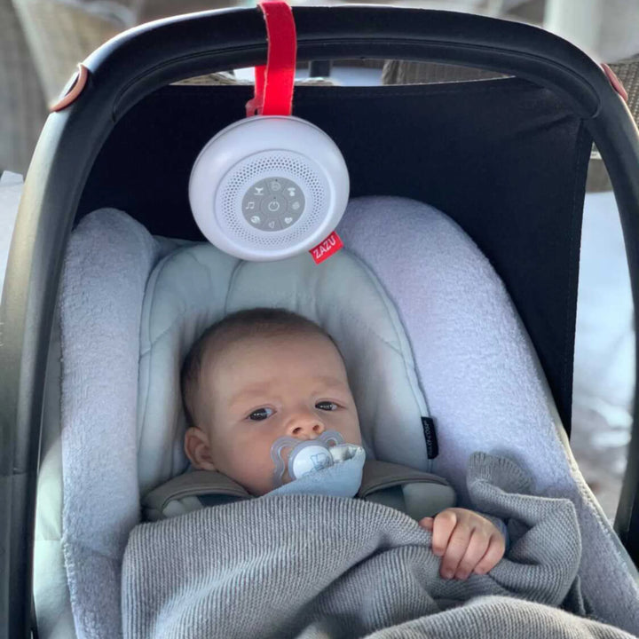Baby sitting calmly in a car seat with a pacifier, soothed by the ZAZU Suzy Portable Baby Soother attached above with a red strap. The white noise machine plays gentle sounds to help babies relax and sleep while traveling. Ideal portable baby sleep aid for car journeys, prams, and on-the-go soothing.